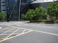 a paved road with many yellow stripes near a tall glass building and trees in the city