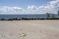 view of a city skyline across the water, with a cobble stone path next to the river, and bench in foreground