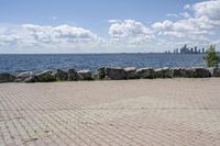 view of a city skyline across the water, with a cobble stone path next to the river, and bench in foreground