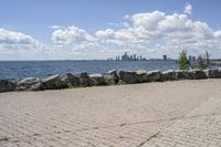 view of a city skyline across the water, with a cobble stone path next to the river, and bench in foreground