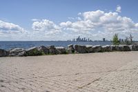 view of a city skyline across the water, with a cobble stone path next to the river, and bench in foreground