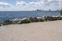 view of a city skyline across the water, with a cobble stone path next to the river, and bench in foreground