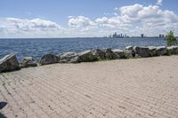 view of a city skyline across the water, with a cobble stone path next to the river, and bench in foreground