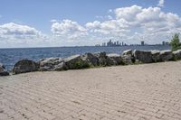 view of a city skyline across the water, with a cobble stone path next to the river, and bench in foreground