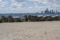 view of a city skyline across the water, with a cobble stone path next to the river, and bench in foreground