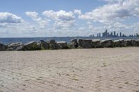 view of a city skyline across the water, with a cobble stone path next to the river, and bench in foreground