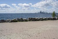 view of a city skyline across the water, with a cobble stone path next to the river, and bench in foreground