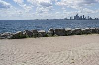 view of a city skyline across the water, with a cobble stone path next to the river, and bench in foreground