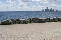 view of a city skyline across the water, with a cobble stone path next to the river, and bench in foreground