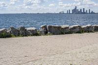 view of a city skyline across the water, with a cobble stone path next to the river, and bench in foreground