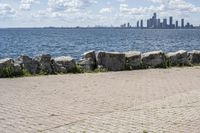 view of a city skyline across the water, with a cobble stone path next to the river, and bench in foreground