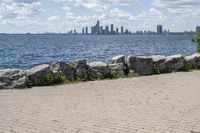 view of a city skyline across the water, with a cobble stone path next to the river, and bench in foreground