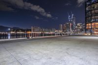 an empty parking lot with several trees near a large body of water at night with skyscrapers in the background