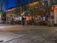 people are walking and walking down the street at night time at a city square, surrounded by trees