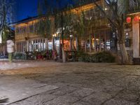 people are walking and walking down the street at night time at a city square, surrounded by trees