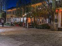 people are walking and walking down the street at night time at a city square, surrounded by trees