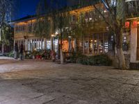 people are walking and walking down the street at night time at a city square, surrounded by trees