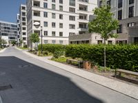 an empty city street between two high rise apartment buildings with plants lining either side and a park bench next to the road