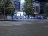 an empty city street with many light posts lit up at night in the background, there is trees growing outside and a building in the center
