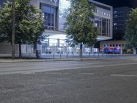 an empty city street with many light posts lit up at night in the background, there is trees growing outside and a building in the center