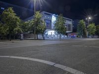 an empty city street with many light posts lit up at night in the background, there is trees growing outside and a building in the center