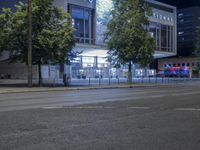 an empty city street with many light posts lit up at night in the background, there is trees growing outside and a building in the center