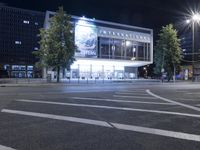 an empty city street with many light posts lit up at night in the background, there is trees growing outside and a building in the center