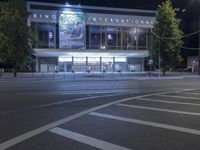 an empty city street with many light posts lit up at night in the background, there is trees growing outside and a building in the center