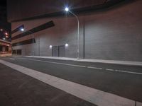 the empty road has two street lamps on it at night, near a brick building