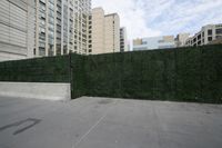 a view of a road through tall buildings with green hedges next to the curb and a gate