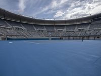 a empty tennis court with rows of empty seats behind it in a city setting with a cloudy sky above the tennis court