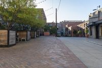 an empty brick sidewalk in front of a red brick building with several chairs, benches and tables on the side