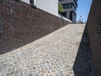 an alley way with brick pavement between the brick walls and building on the left, and a side walk to another