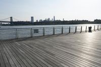 a wide wooden boardwalk with the water in the background and a city behind it and the sun shining