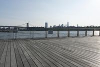 a wide wooden boardwalk with the water in the background and a city behind it and the sun shining