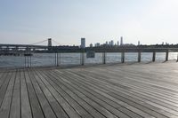a wide wooden boardwalk with the water in the background and a city behind it and the sun shining