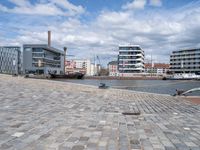 the road is paved with gray cobbles next to buildings and boats, with a bench at the edge