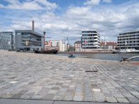 the road is paved with gray cobbles next to buildings and boats, with a bench at the edge