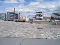 the road is paved with gray cobbles next to buildings and boats, with a bench at the edge