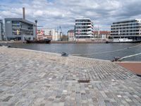 the road is paved with gray cobbles next to buildings and boats, with a bench at the edge