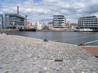 the road is paved with gray cobbles next to buildings and boats, with a bench at the edge