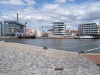 the road is paved with gray cobbles next to buildings and boats, with a bench at the edge