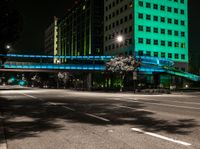 a bright lit up pedestrian bridge over an empty road at night in the city with cars going by