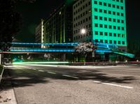 a bright lit up pedestrian bridge over an empty road at night in the city with cars going by