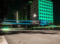 a bright lit up pedestrian bridge over an empty road at night in the city with cars going by