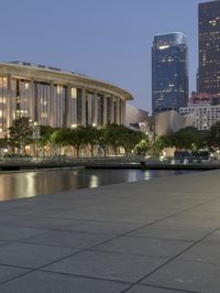 the walkway at the park leads to a lake and buildings in the background as evening falls