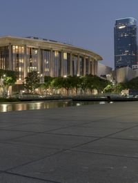 the walkway at the park leads to a lake and buildings in the background as evening falls