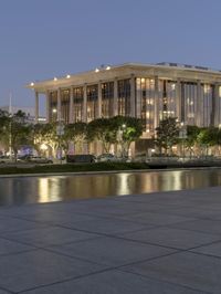 the walkway at the park leads to a lake and buildings in the background as evening falls