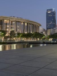 the walkway at the park leads to a lake and buildings in the background as evening falls