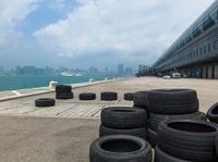 many tires have been set up along the walkway on a dock by a harbor with a cityscape in the background